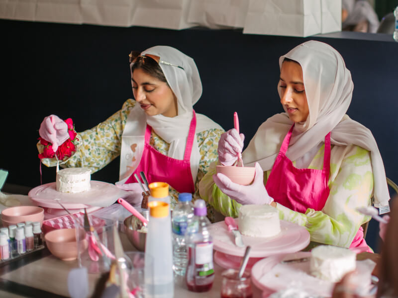 Two women in hijabs and pink aprons icing cakes